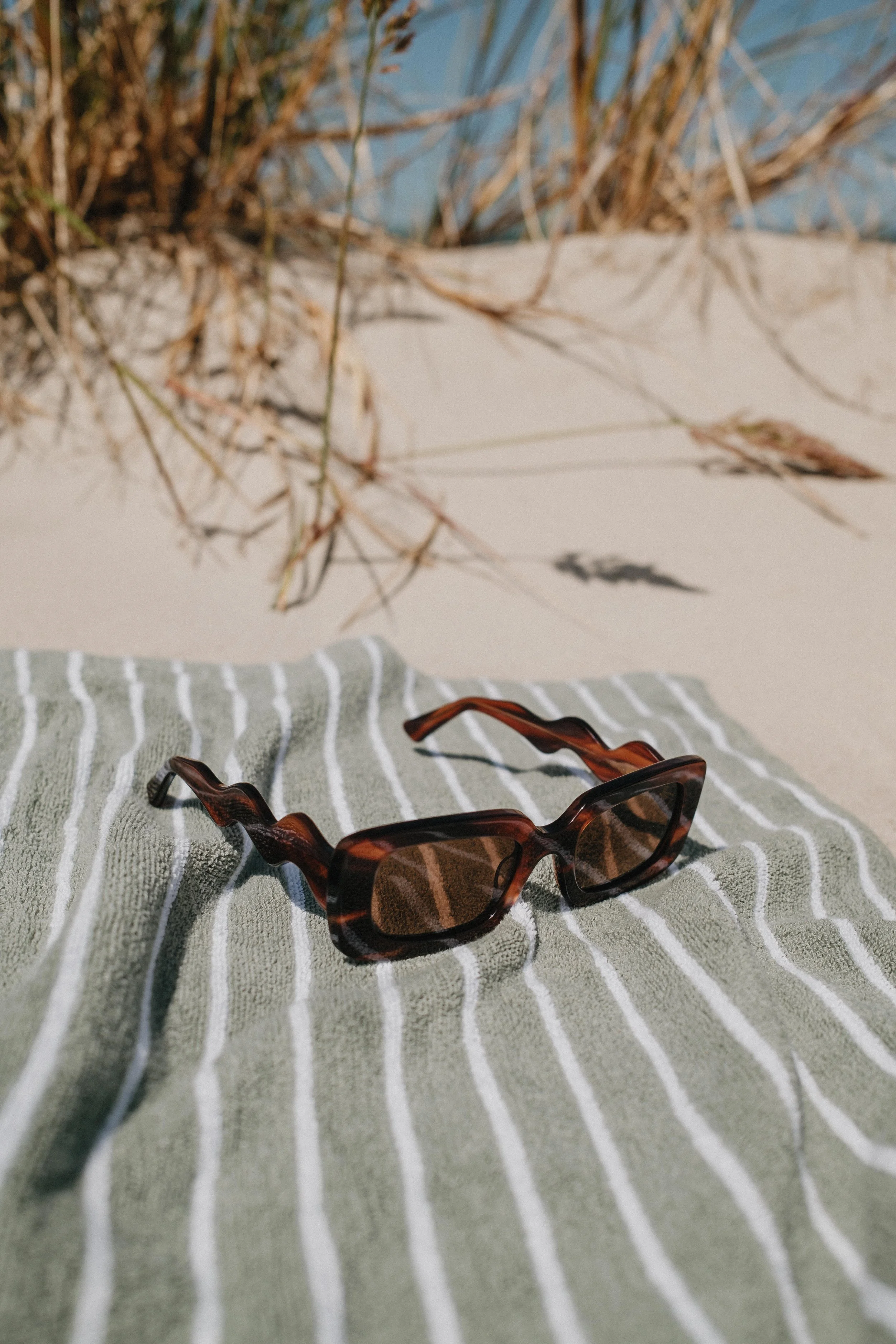 Brown tortoiseshell sunglasses resting on a gray and white striped fabric, with dried grass and a blue sky reflection in the background.