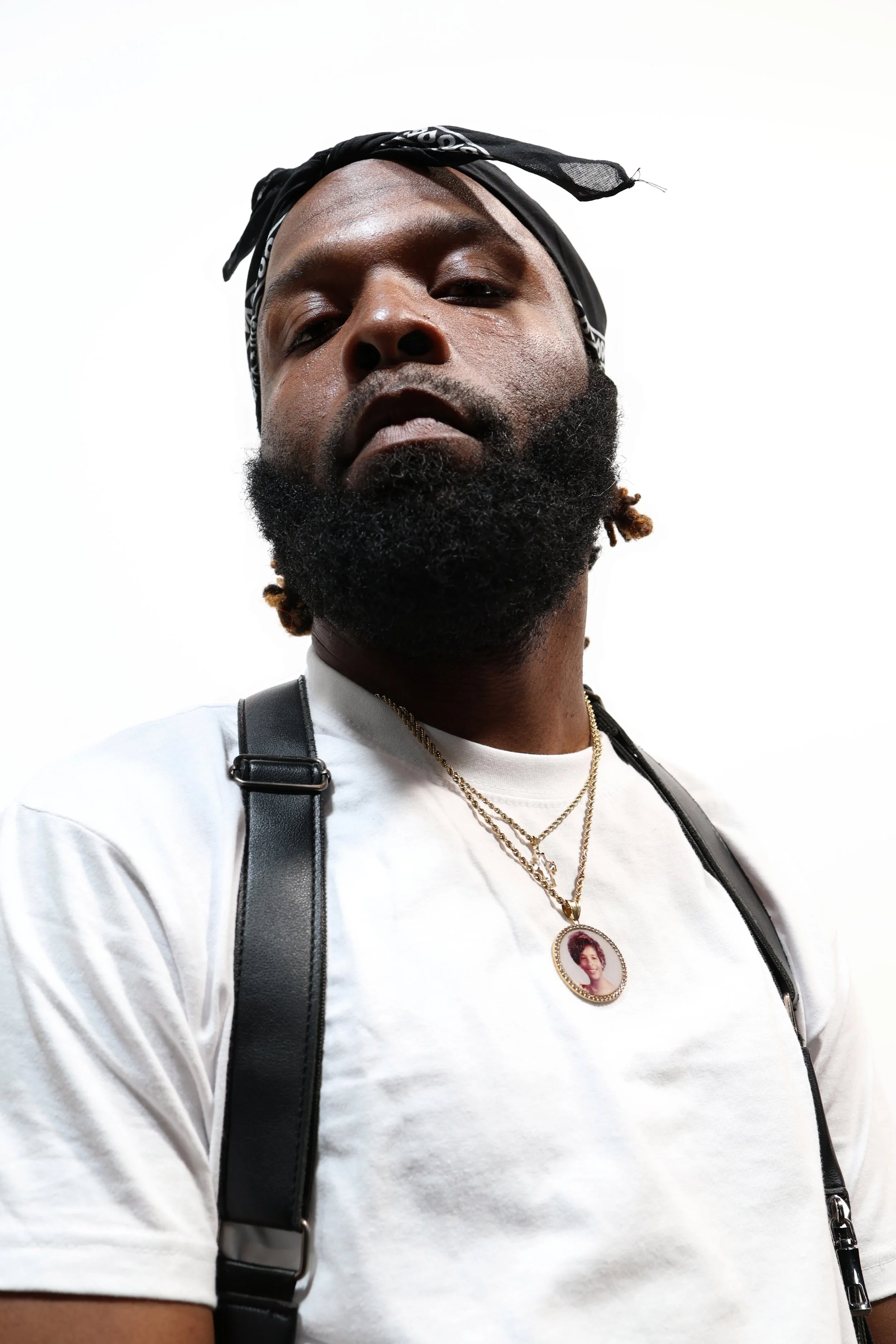 Close-up of a man with a black bandana on his head, gold jewelry, and wearing a white t-shirt. He has a beard and is looking into the camera with a neutral expression. The background is plain white.