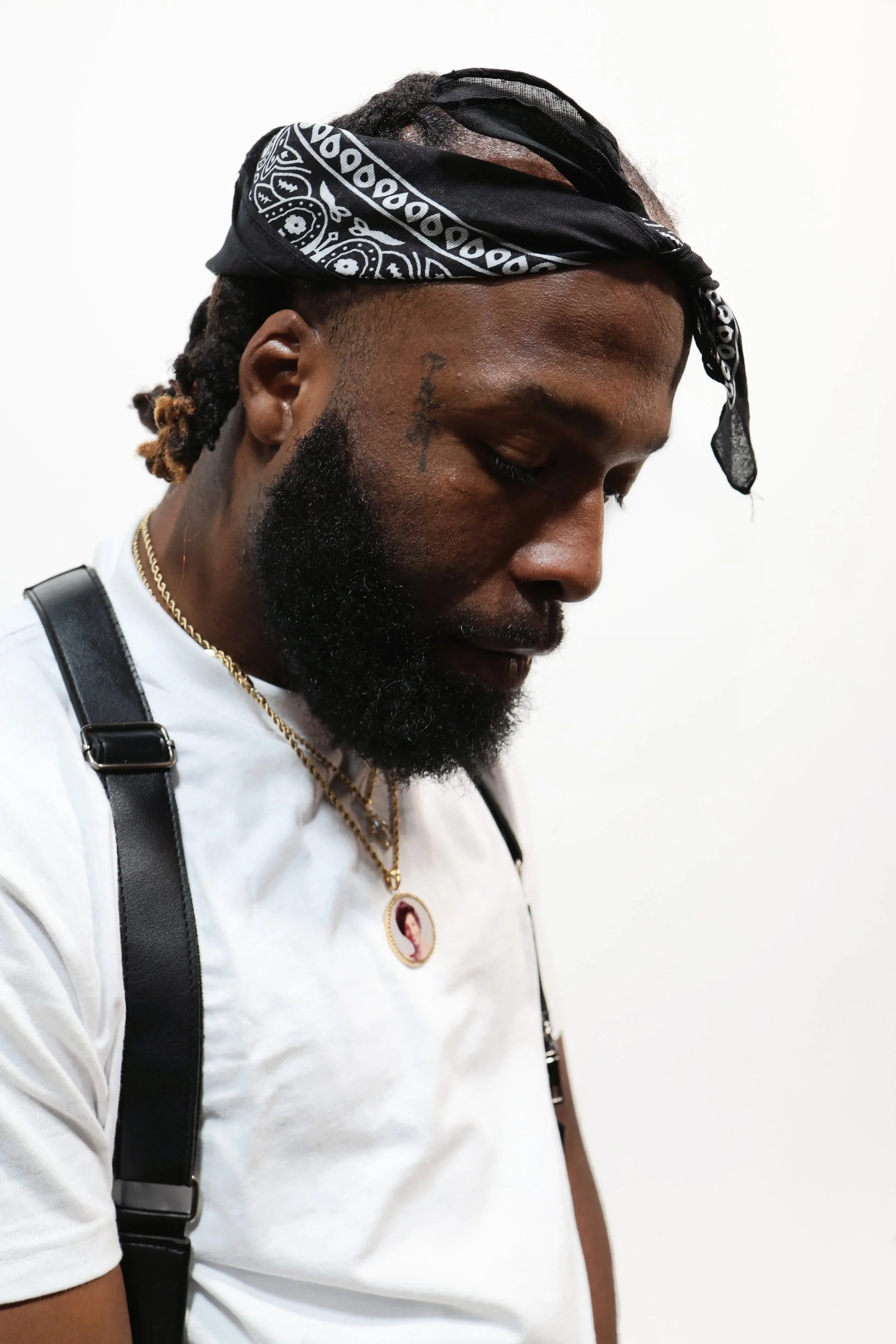 Close-up of a man with a beard and tattoos, wearing a black bandana, gold necklaces, and a white T-shirt with black straps over his shoulders, standing against a plain white background.