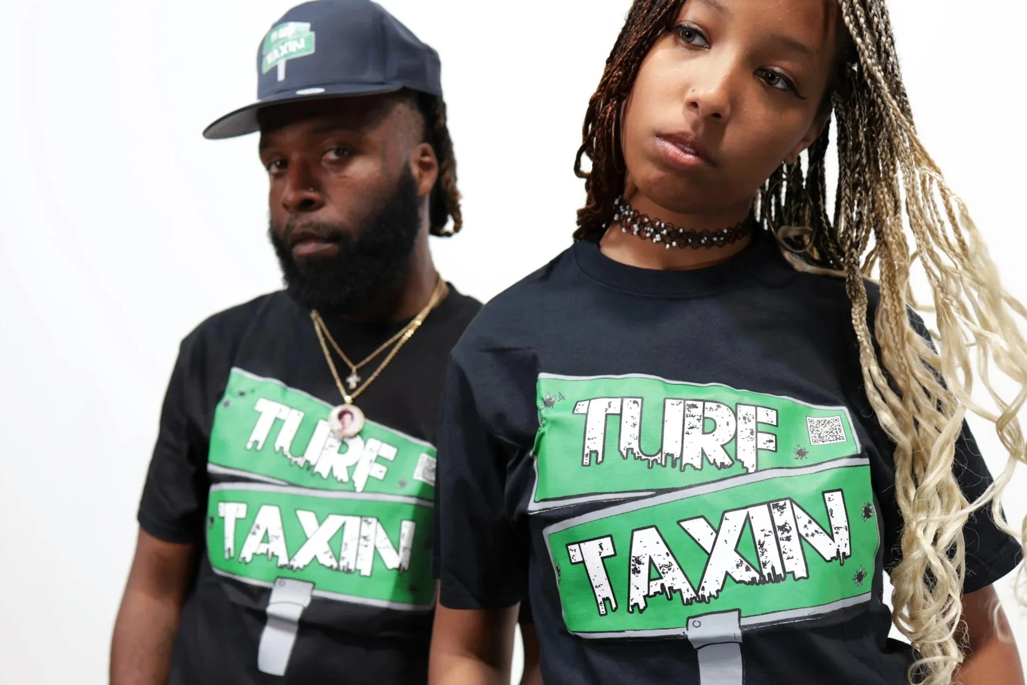 Two people wearing black T-shirts with green and white text that reads 'TAX THE RICH, TAX THE WEALTH'. The person on the right has long twisted hair and is wearing a choker necklace. The person on the left has a beard, a gold chain, and a cap.