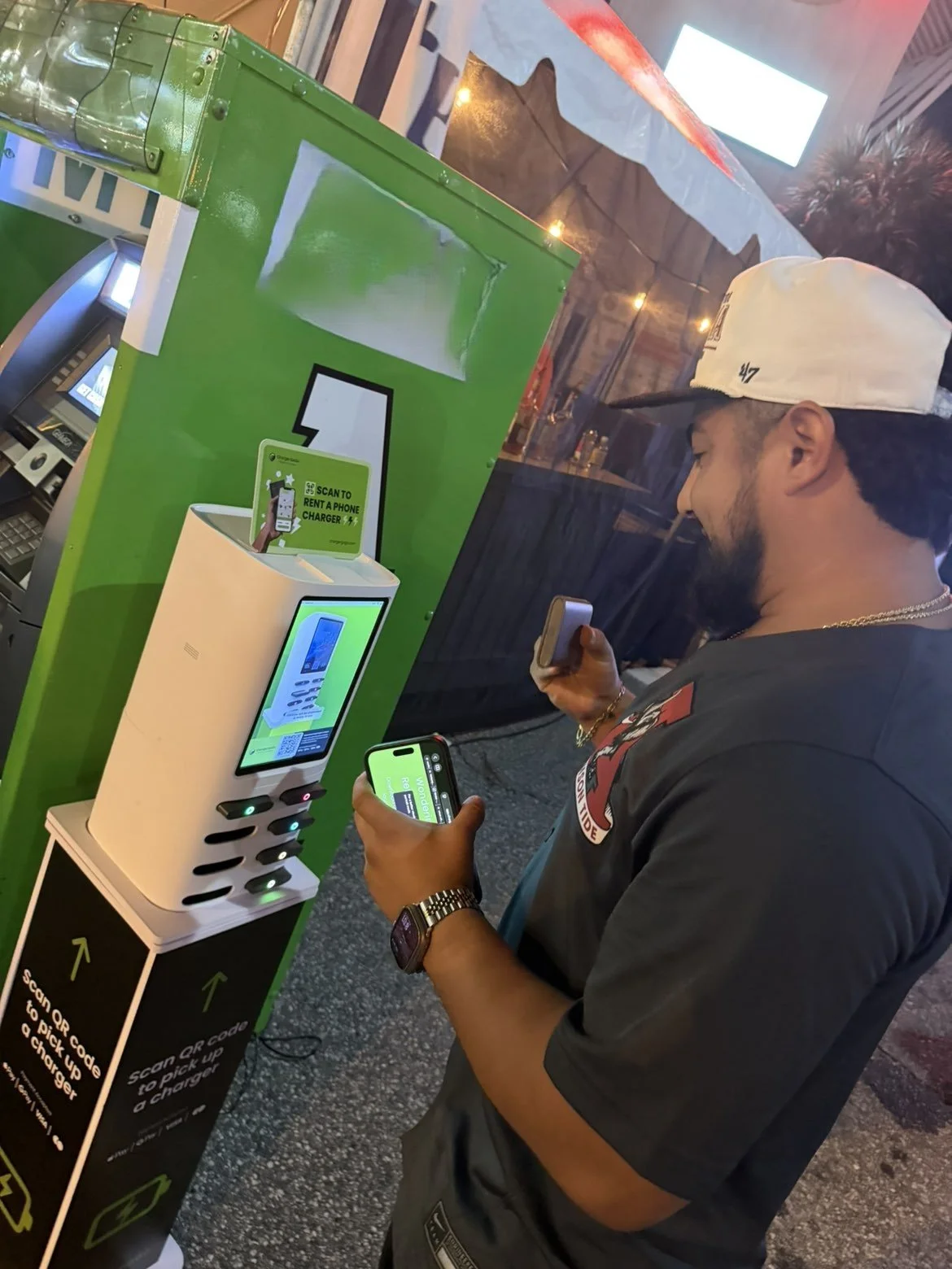 A man at a green and white contactless phone charging station, scanning QR codes with his smartphone and a small device, with a sign instructing to scan a QR code to pick up a charger at the station.