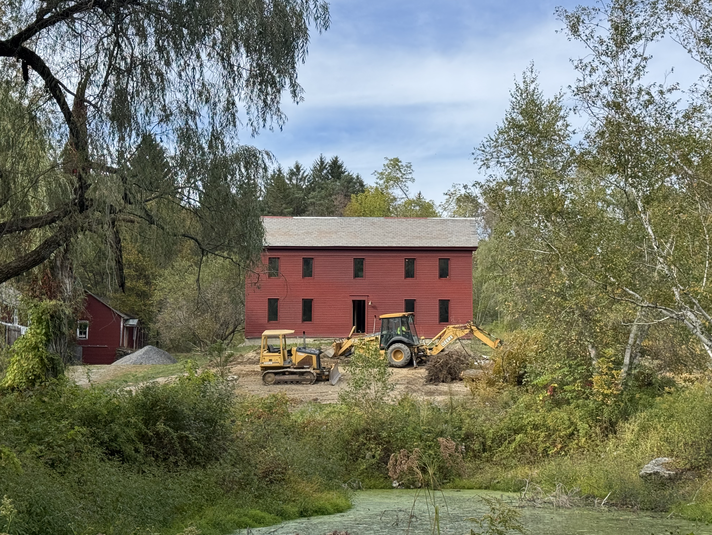 A construction site with a yellow backhoe loader on dirt, a red building with black windows in the background, and trees with green foliage surrounding the area under a partly cloudy sky.