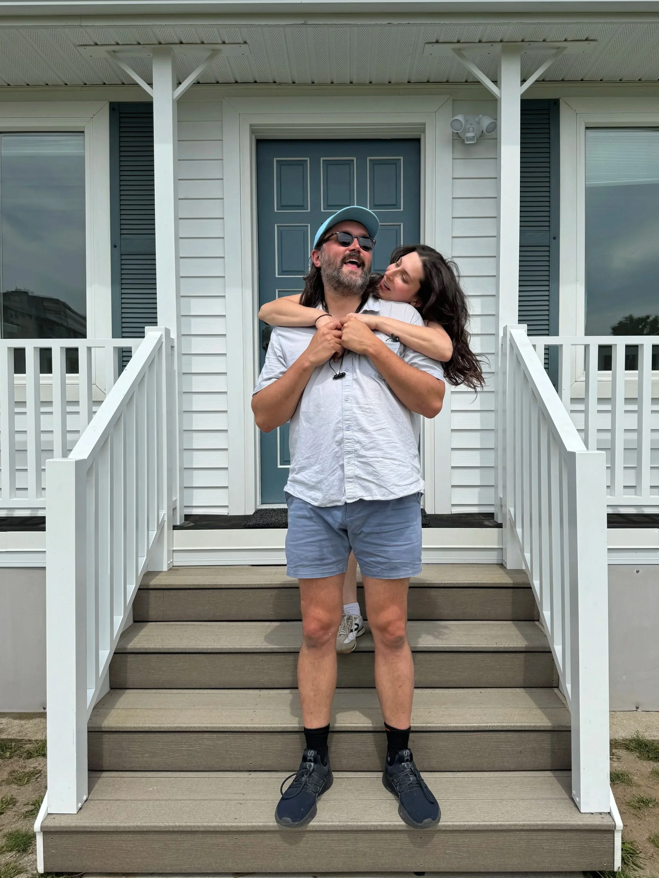A man and a woman standing on a porch in front of a blue door. The woman is hugging the man from behind, and they are both smiling. The man is wearing sunglasses, a baseball cap, a white shirt, shorts, and running shoes. The woman has long dark hair, and they are casually dressed.