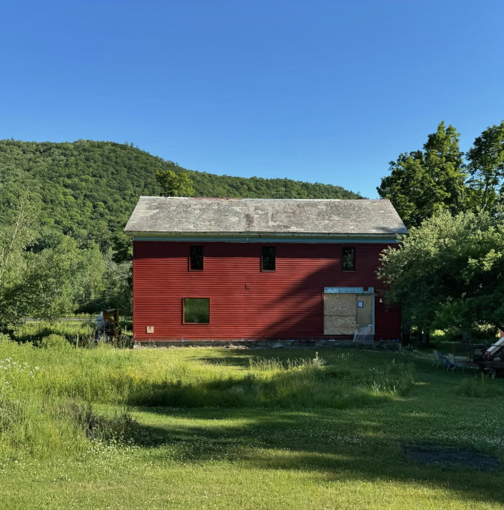 A red two-story house with several windows, surrounded by lush green grass and trees, with a backdrop of green hills and a clear blue sky.