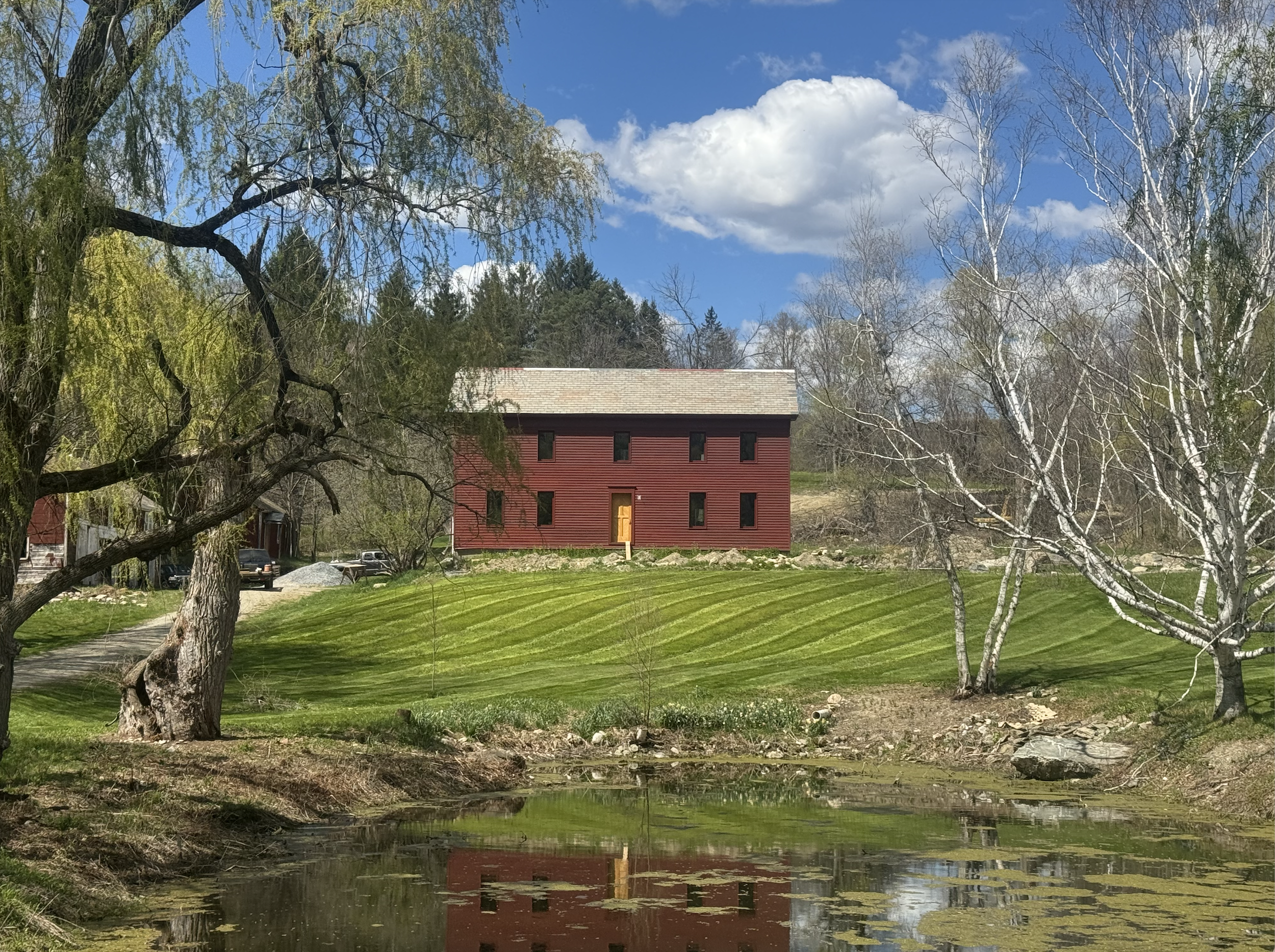 A red house with black trim in a grassy area surrounded by leafless trees, with a small pond in the foreground reflecting the house and trees, under a partly cloudy blue sky.