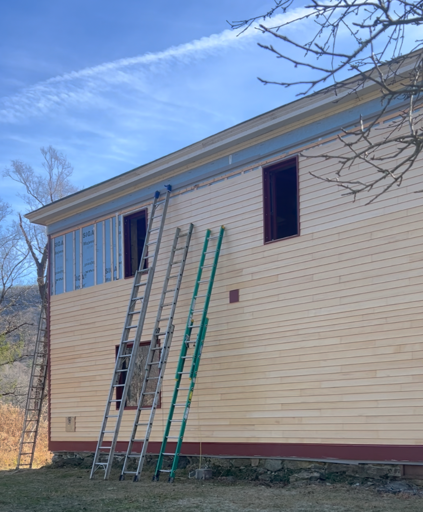 Side of a house under construction with several ladders leaning against it, showing a partially completed exterior with wooden siding and two open window spaces.