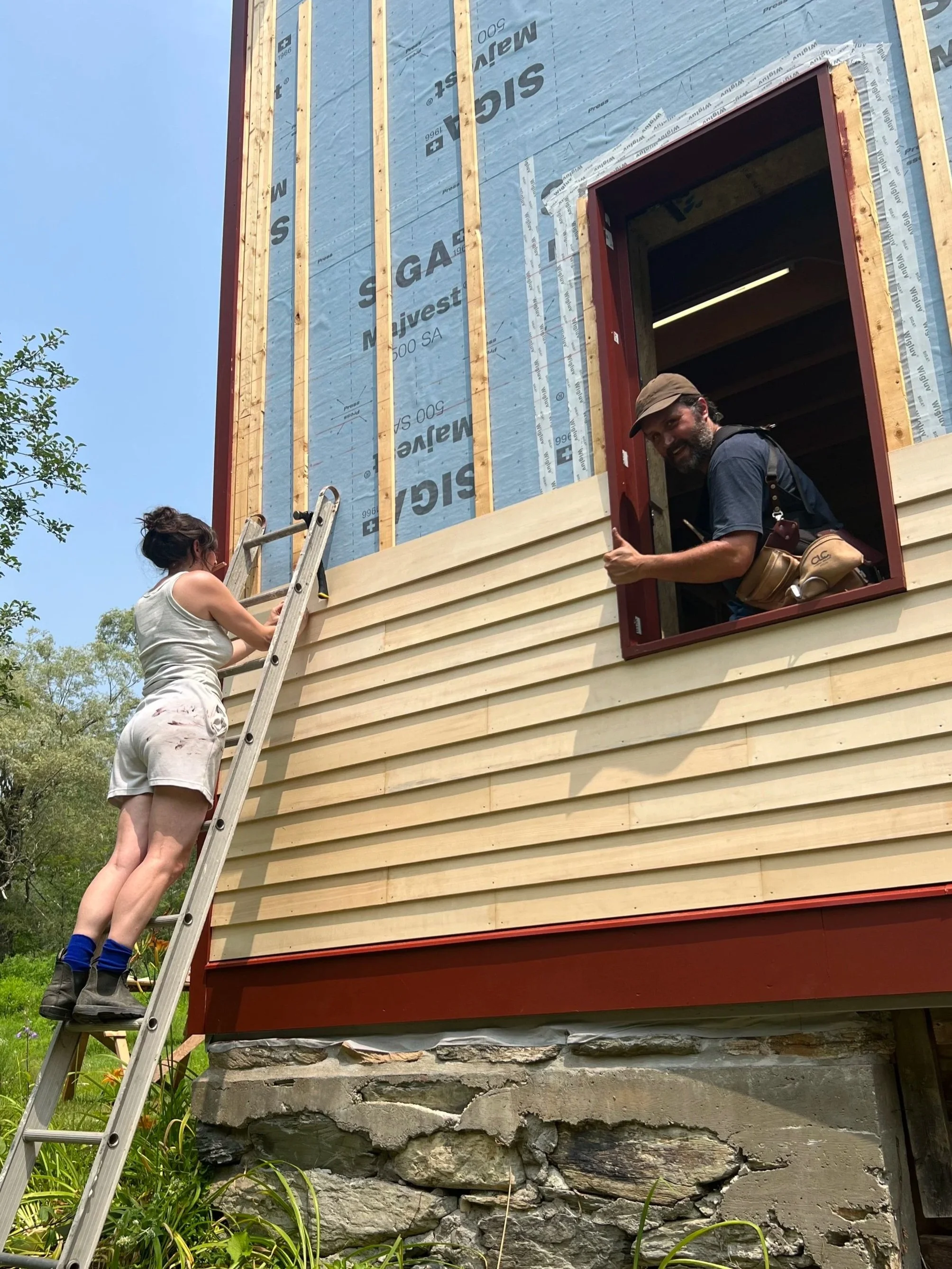 Two people working on the construction of a house. The woman is on a ladder, holding a tool, and the man is inside the house looking out of a window, smiling.