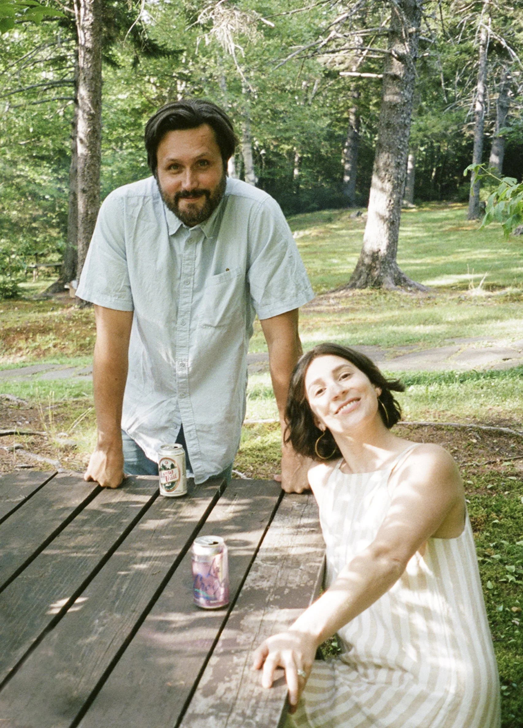 A man and a woman outdoors at a park or forest area, with trees and greenery in the background. The man is standing behind a wooden picnic table, leaning forward slightly, with dark hair, a beard, wearing a light-colored shirt. The woman is sitting at the table, smiling, with dark hair, wearing a light-colored, striped dress. There are cans of drinks on the table, and sunlight filters through the trees.