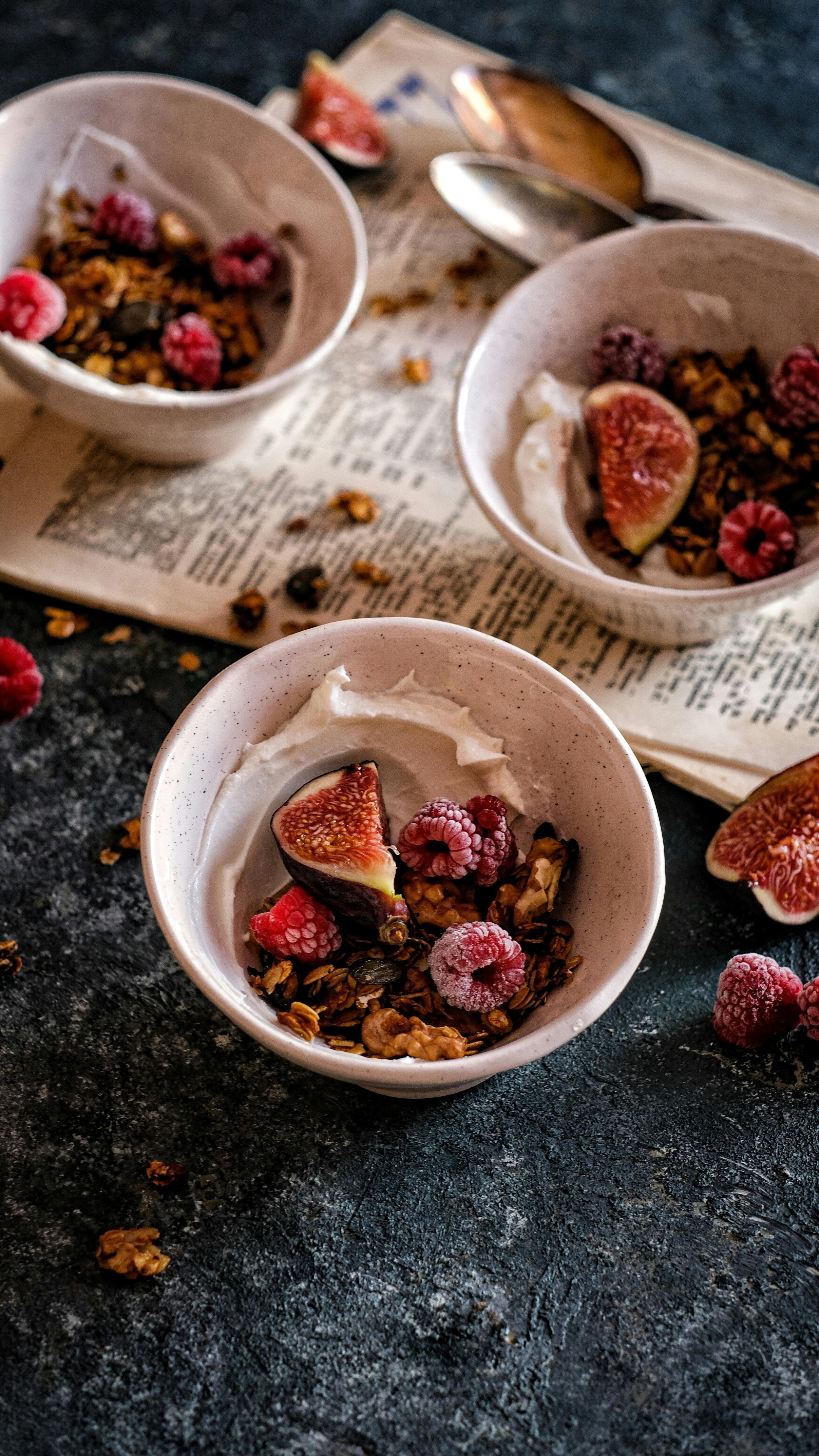 private chef las vegas nv. Close-up of three white bowls filled with yogurt topped with granola, fresh figs, and raspberries, on a dark textured surface with scattered raspberries and granola, and vintage spoons.