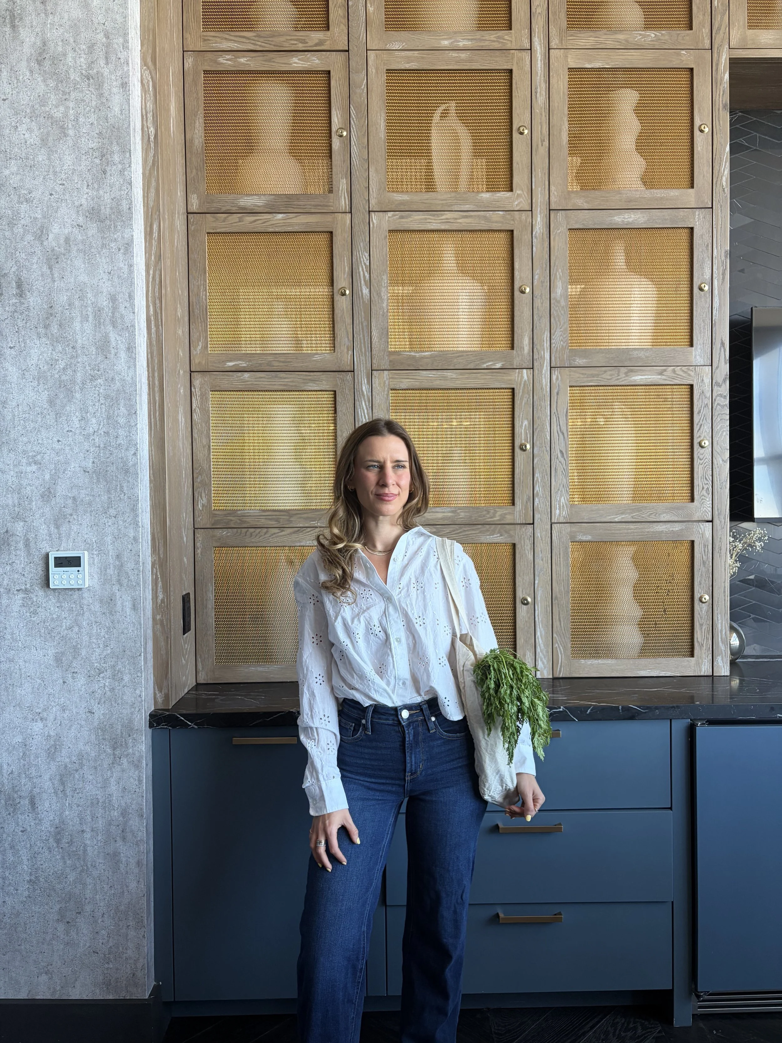 Nutritionist las vegas standing in front of a wooden cabinet with glass pane doors, holding a tote bag with green leafy vegetables, in a modern kitchen.