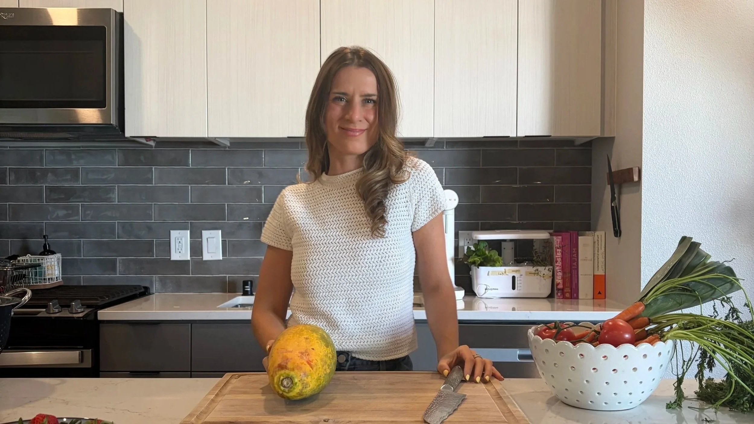 Woman with brown hair and a white knit top standing in a modern kitchen, with a papaya on the cutting board, a knife, and a bowl of tomatoes and vegetables on the counter.