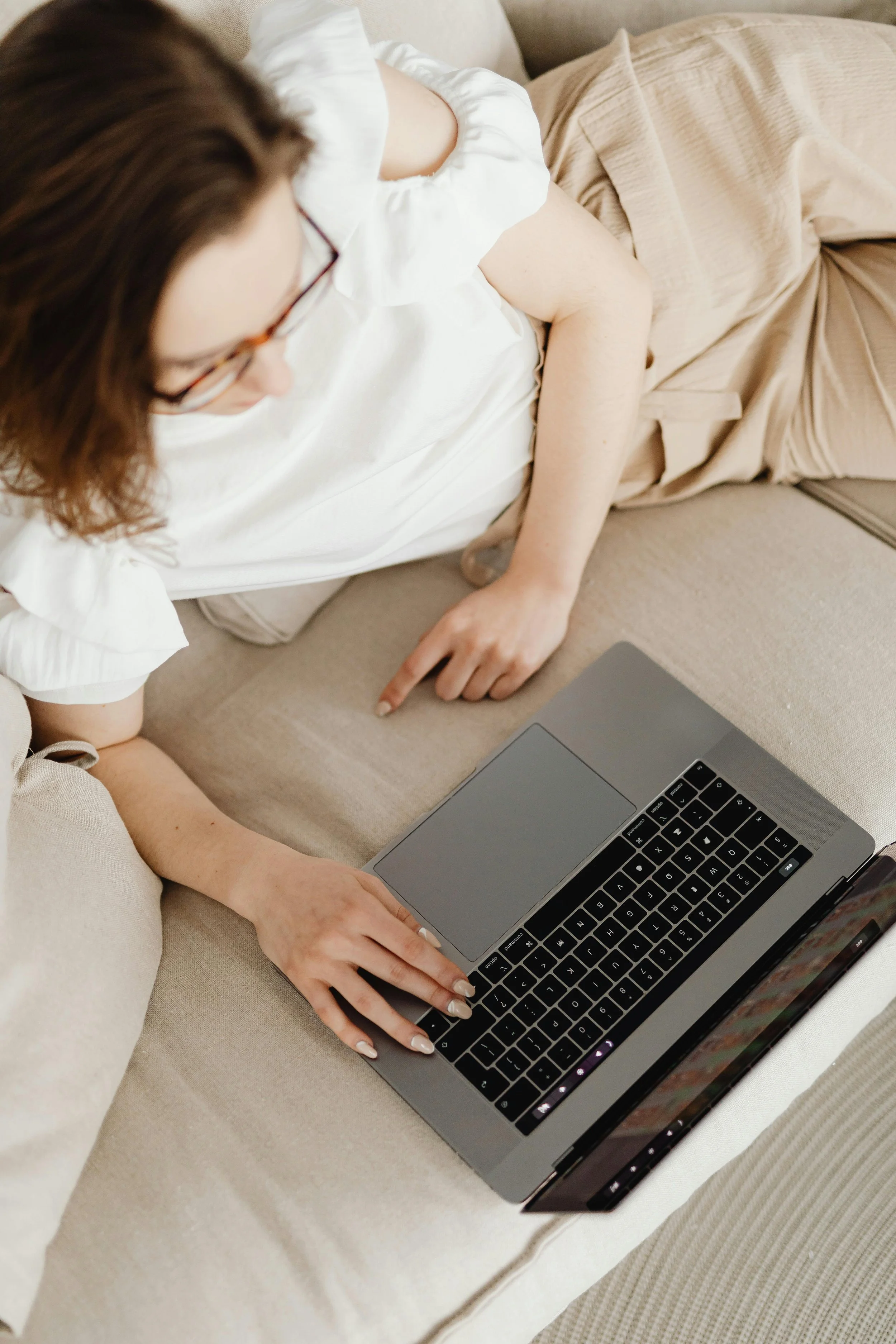 A woman with glasses lying on a couch, pointing at a silver laptop placed on the couch.