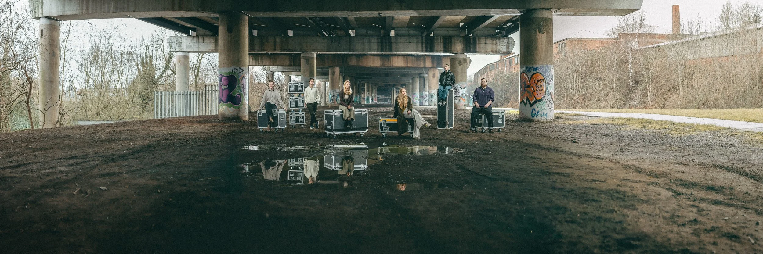 A group of six people sitting and standing on flightcases cases under a graffiti-covered overpass under spagetti junction