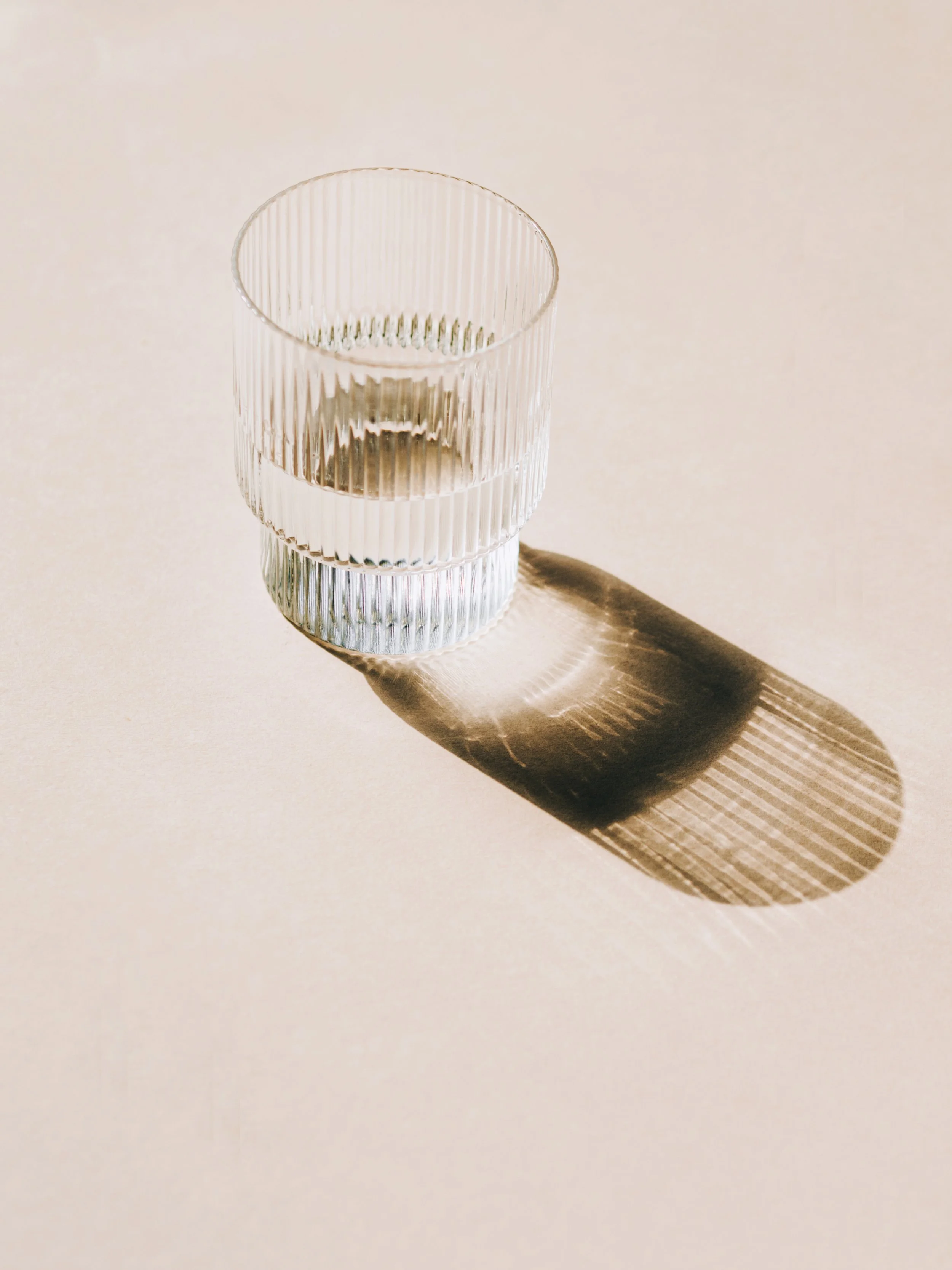 A clear glass with vertical ridges filled with water, casting a shadow on a plain light-colored surface.