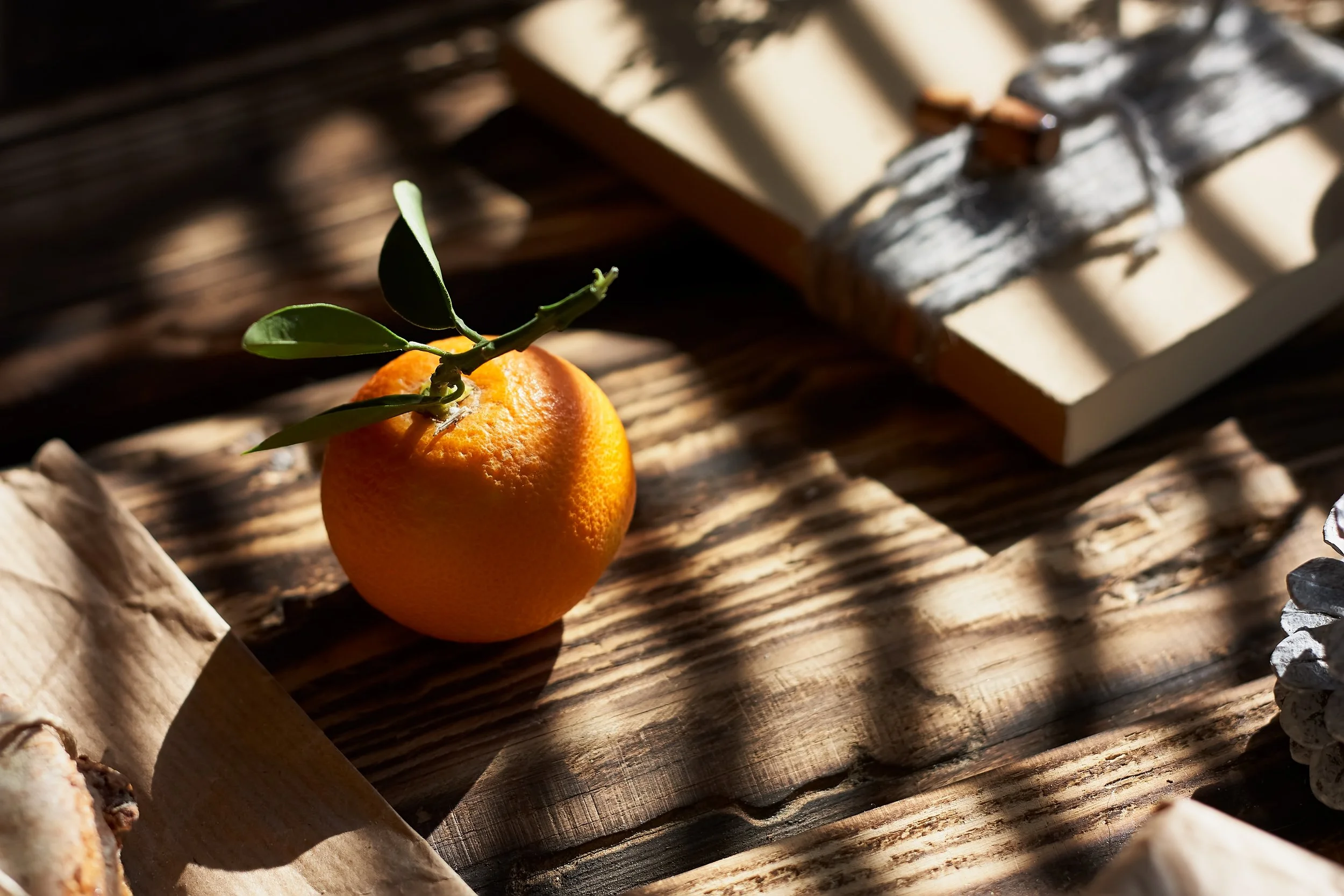 A close-up of an orange with green leaves attached, resting on a rustic wooden surface, with sunlight creating shadows.