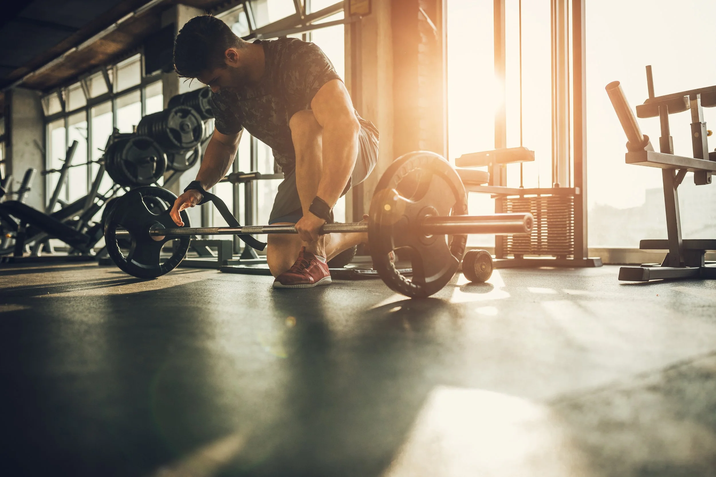 A man in athletic wear is preparing to lift a barbell on the floor in a gym with large windows and sunlight streaming in.