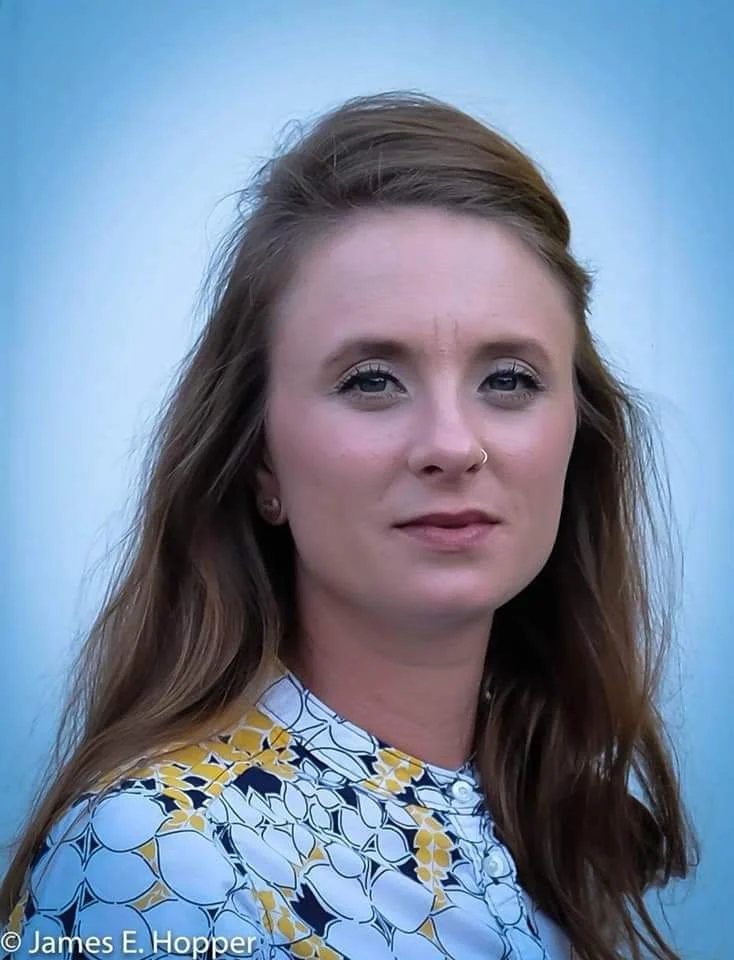 A woman with long brown hair, wearing a white, yellow, and black patterned top, standing outdoors against a blue sky background.