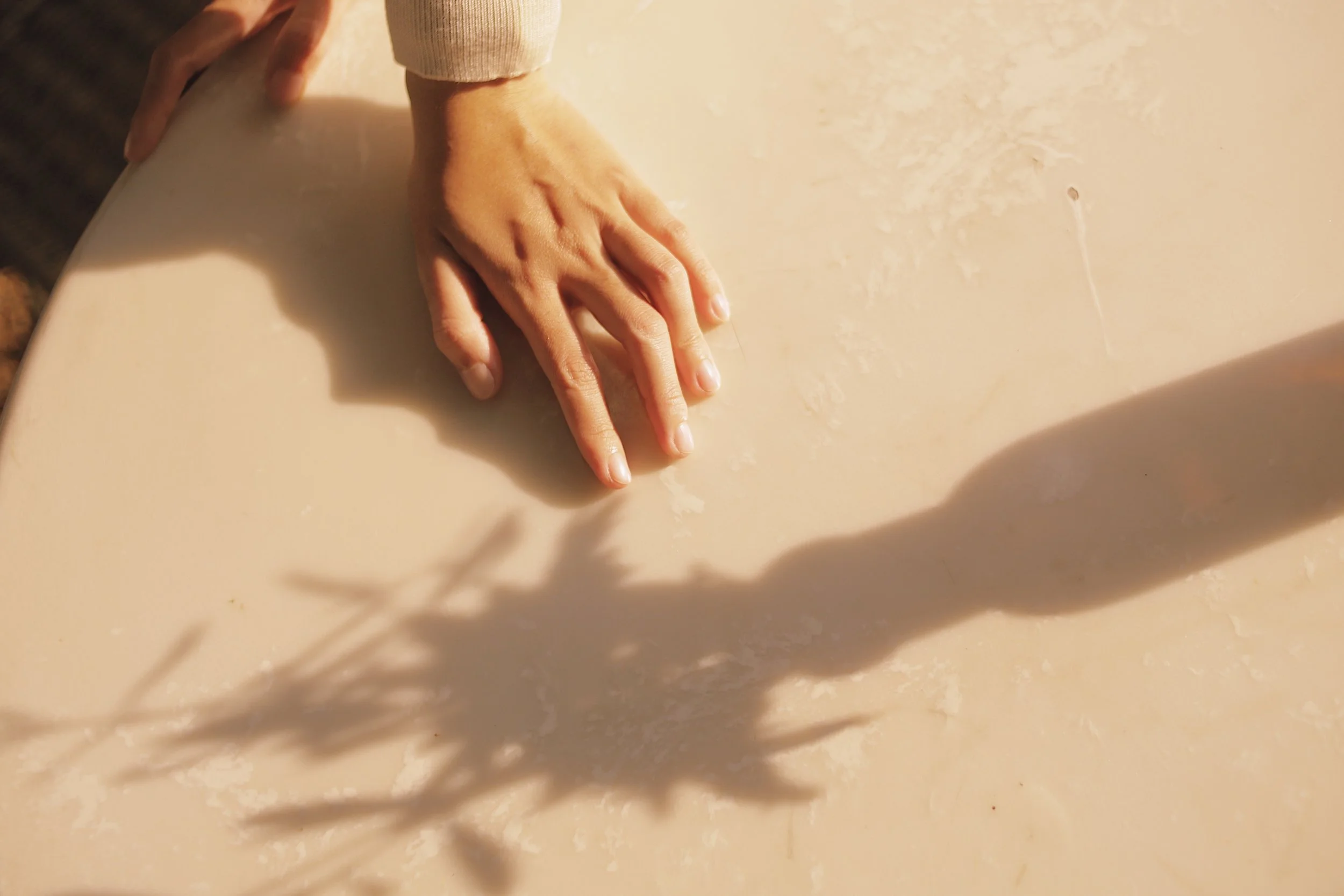A person's hand with a light skin tone resting on a beige marble or stone surface, with the person wearing a beige or light-colored long-sleeve shirt.