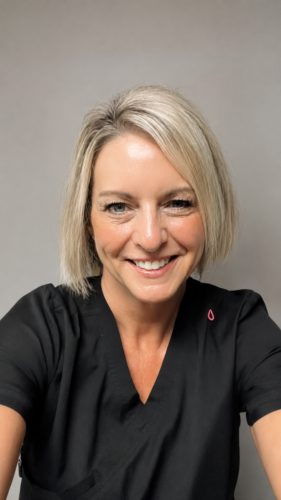 A woman with blonde, shoulder-length hair smiling in a black medical scrub top against a plain gray background.