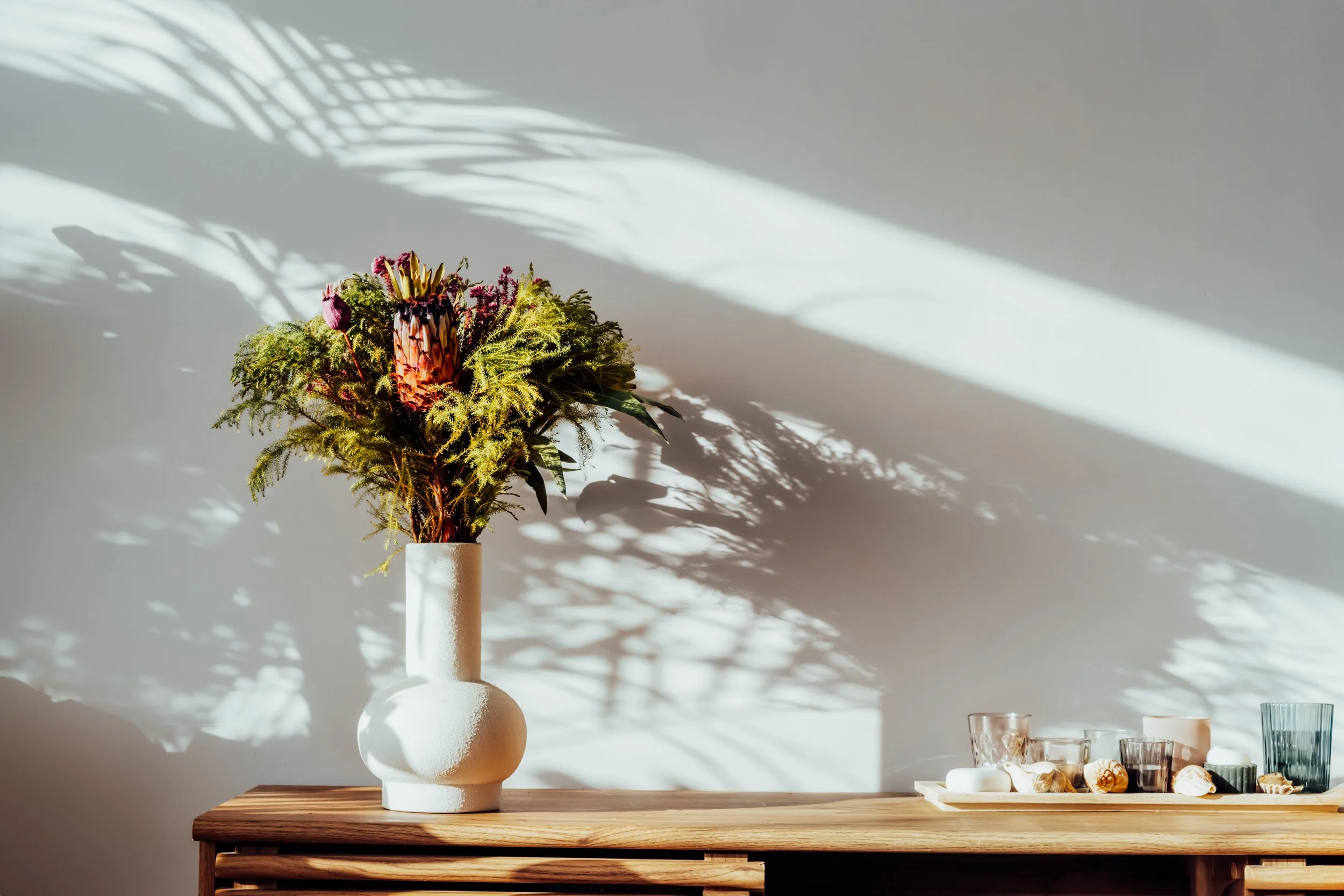 A tall bouquet of assorted flowers and greenery in a textured white vase on a wooden table, with sunlight and shadow patterns on the wall behind.