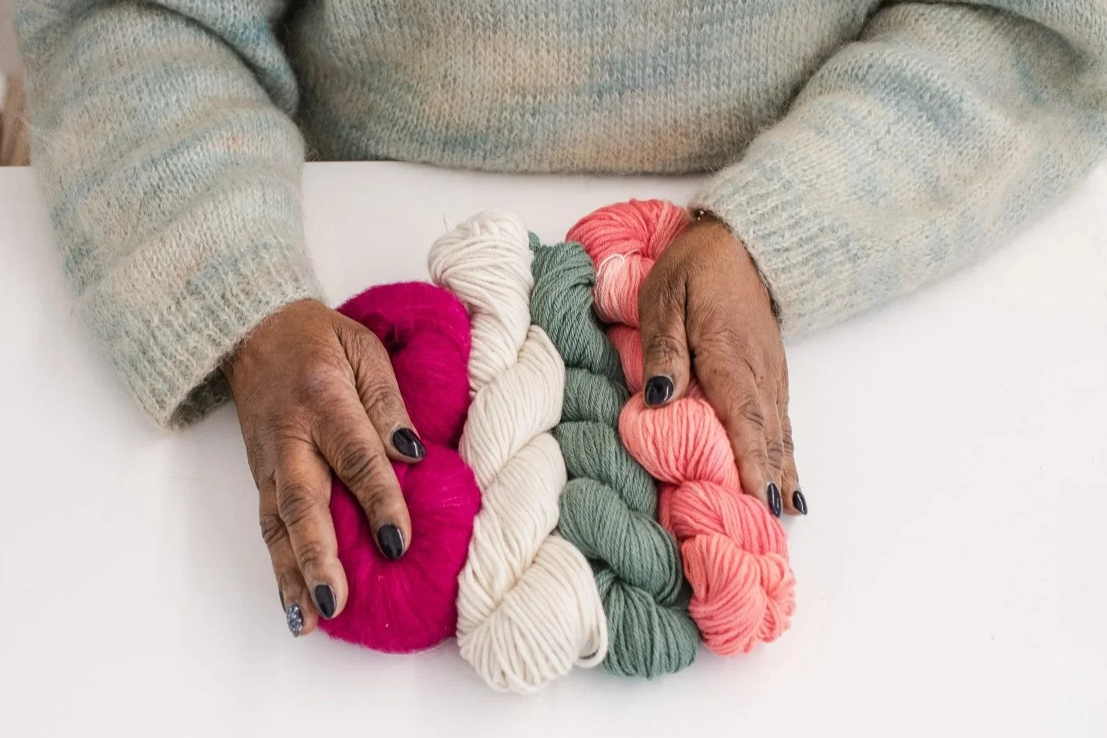 A person's hands holding five balls of yarn in different colors: pink, white, green, and peach, on a white surface. The person is wearing a light gray knitted sweater.