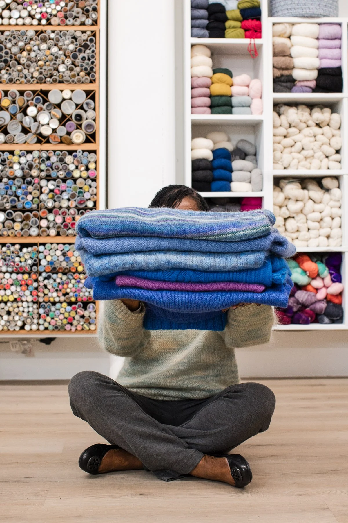 Person sitting cross-legged holding a large stack of folded sweaters in front of their face in a yarn store, with shelves of yarn and fabric in the background.