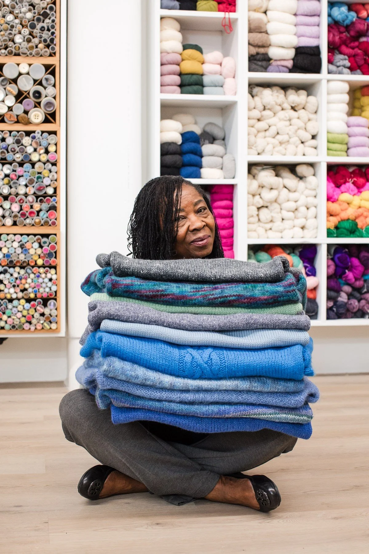 A woman sitting on the floor partially surrounded by a large stack of folded sweaters in various shades of blue, gray, and green. Behind her are shelves filled with colorful yarn or fabric supplies.