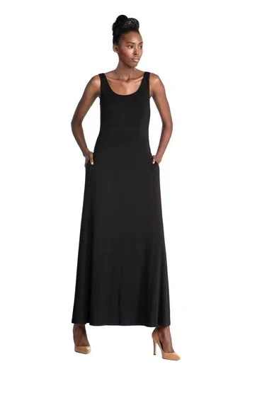A woman in a long black sleeveless dress with a scoop neckline standing with her hands in her pockets against a white background.