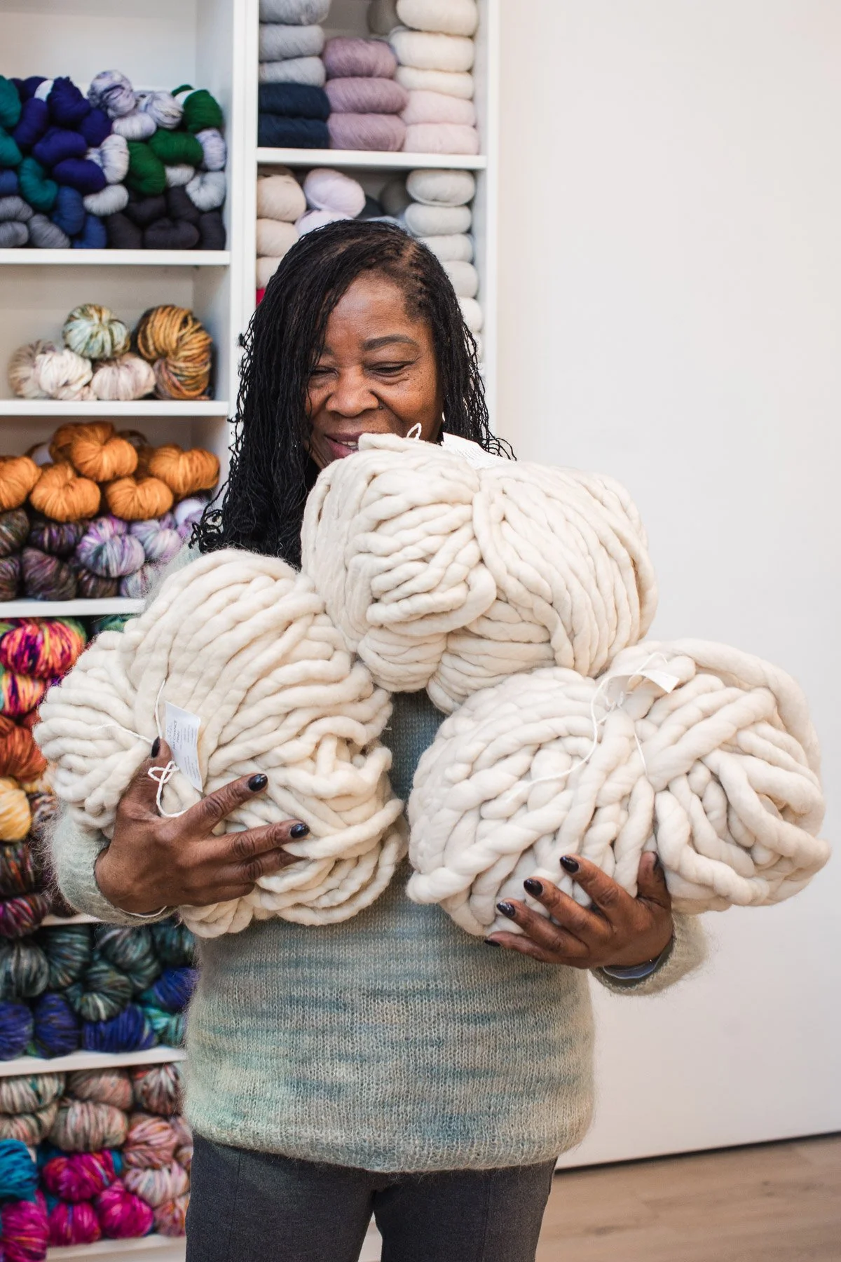 A woman holding several large rolls of thick, chunky white yarn in a craft store. Behind her, shelves display colorful skeins of yarn.