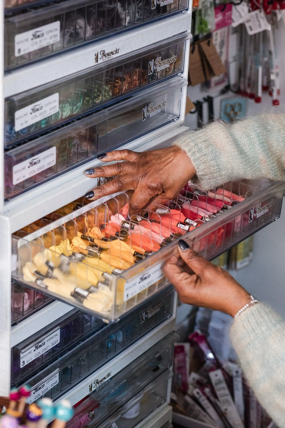 Close-up of a person's hand reaching into a plastic drawer filled with colorful embroidery floss or threads in a craft store. A second hand is opening another drawer with similar craft threads. The background shows more organized craft supplies and tools.