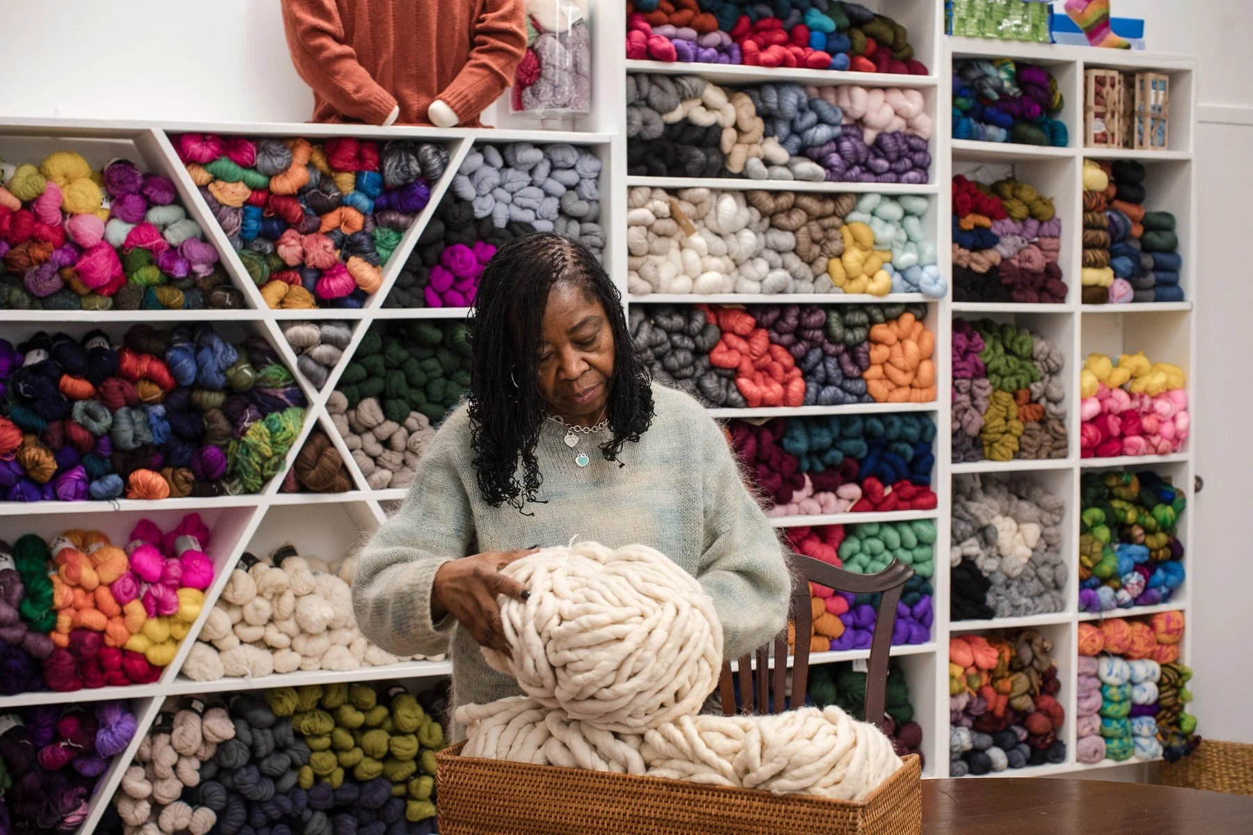 A woman in a gray sweater working with large beige yarn in a craft store, surrounded by shelves filled with colorful yarns.