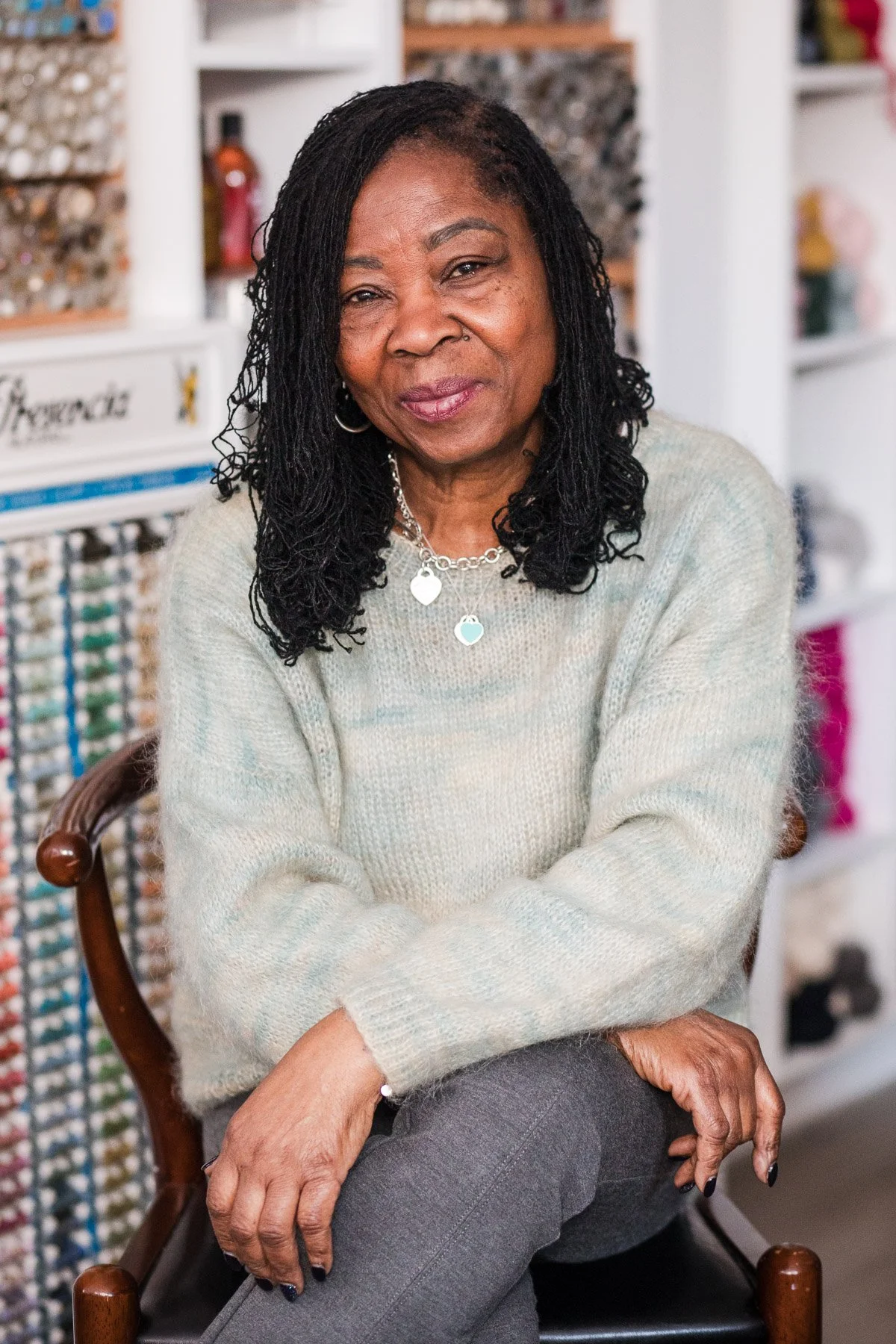 A woman with black, shoulder-length dreadlocks and hoop earrings sitting on a wooden chair, wearing a light-colored sweater with a heart-shaped necklace, in a room with shelves filled with nail polish bottles and beauty products.