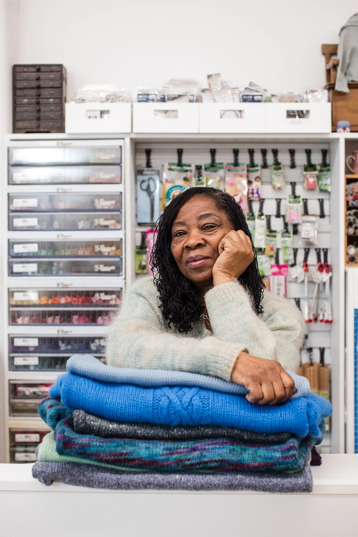 A woman with curly black hair sitting at a table with a stack of folded sweaters in front of her, in a craft or shop setting with shelves of supplies and tools behind her.