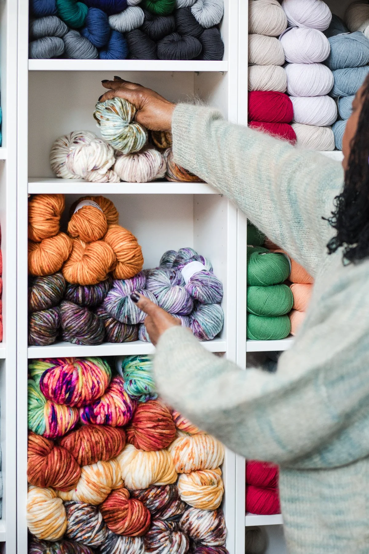 A person in a light-colored sweater is selecting multicolored yarn skeins from a white storage unit filled with various skeins of yarn in different colors, including shades of orange, purple, green, and multicolored patterns.