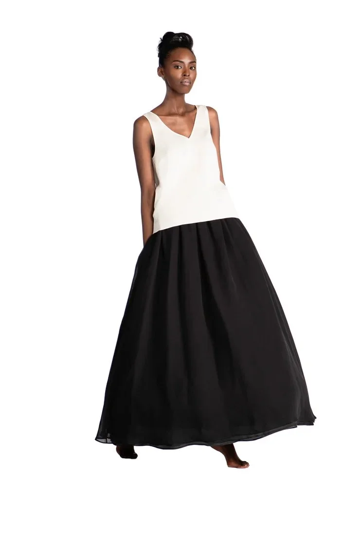 A woman wearing a sleeveless, white top with a black floor-length skirt, standing against a plain white background.