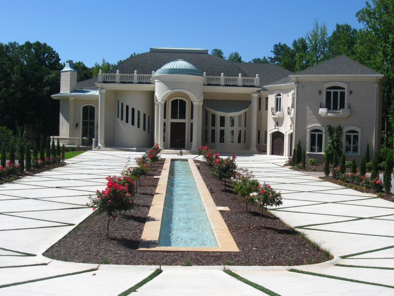 A large, luxurious mansion with a fountain and a landscaped driveway with rose bushes in the front yard.