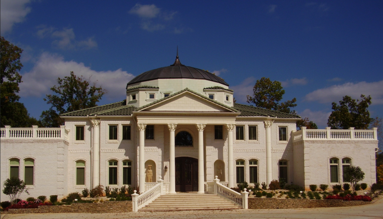Large white mansion with a domed roof, columns at the entrance, surrounded by landscaped garden and trees, under a partly cloudy sky.