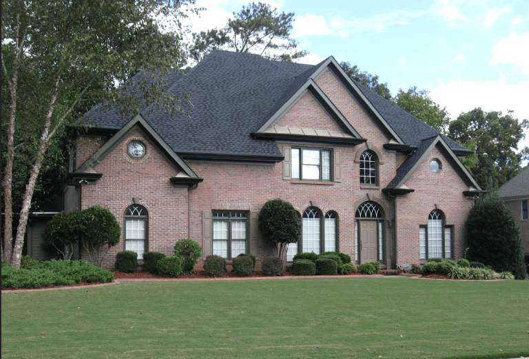 A large brick house with multiple peaked roofs, several windows with shutters, and a small front porch, surrounded by well-maintained green lawn and shrubs.