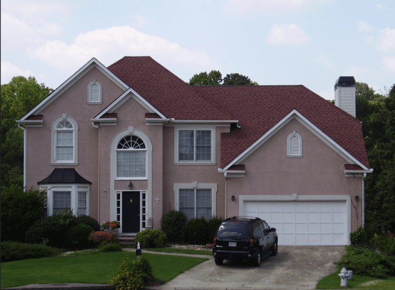 Two-story suburban house with pinkish-beige exterior, red roof, and white trim. Has a black front door, large arched window, garage, and a front lawn with shrubs and flowers. Black SUV parked in driveway.