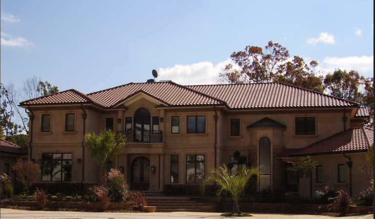 Large two-story house with a red-tiled roof, beige stucco exterior, and landscaped front yard with small palm trees and bushes.