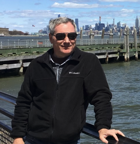 A man wearing sunglasses and a black Columbia jacket stands outdoors near a body of water with a city skyline in the background.