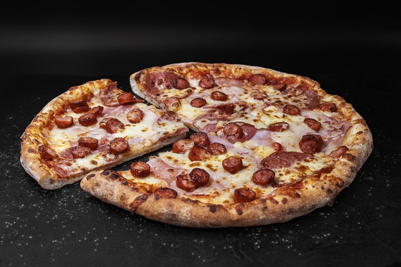 O pizza cu pepperoni și șuncă, din care s-a scos o felie, așezată pe o suprafață neagră.