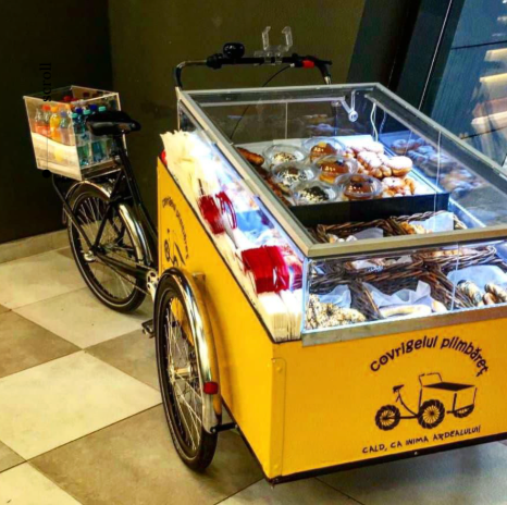 O bicicletă galbenă prevăzută cu o vitrină de sticlă, plină cu gogoși și produse de patiserie, lângă o cutie cu sticle colorate de băuturi.