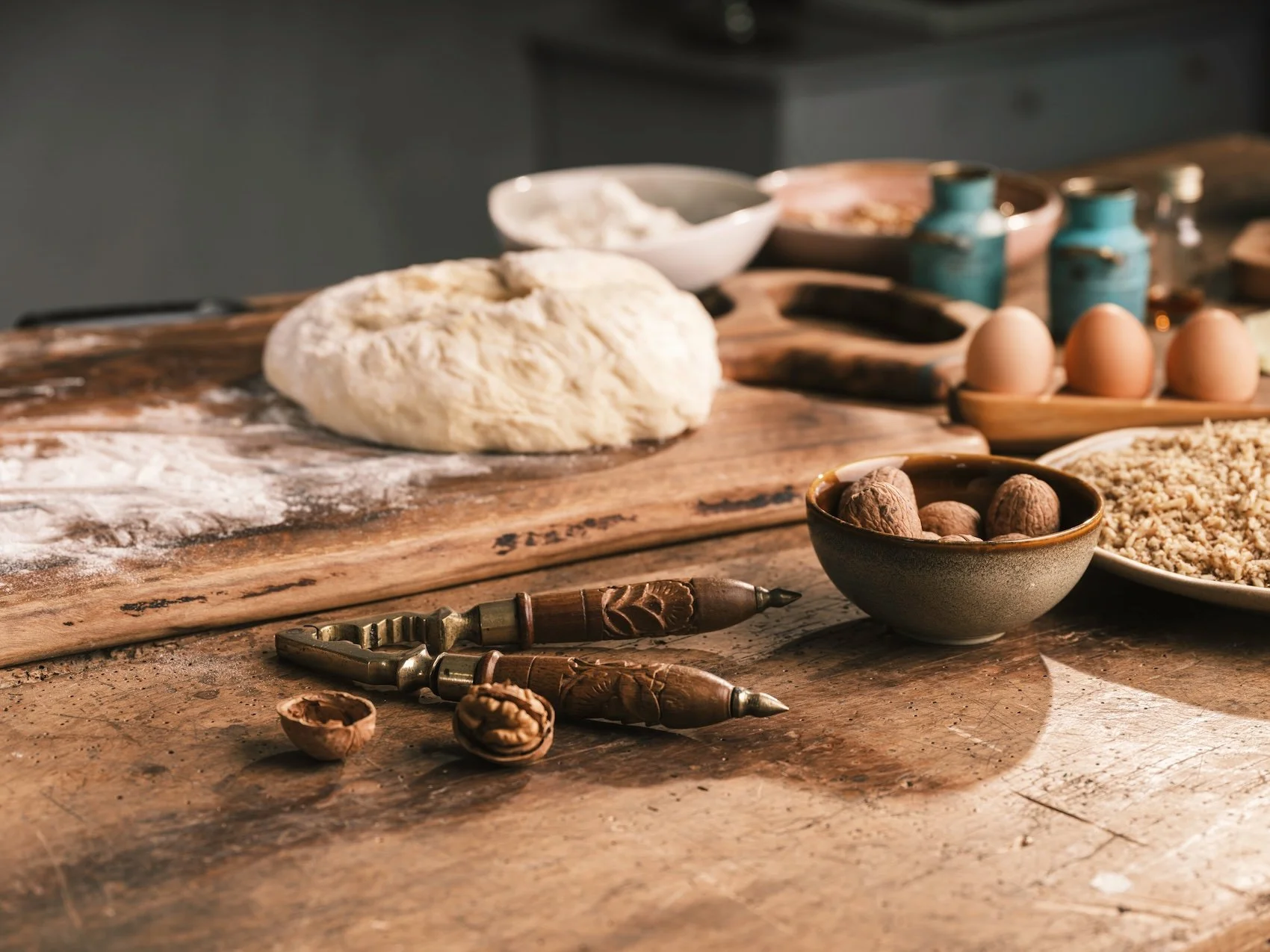 O masă de bucătărie rustică din lemn, pe care se află un ghem de aluat, nuci și ustensile de gătit, înconjurată de ouă, borcane de ceramică și boluri cu ingrediente.