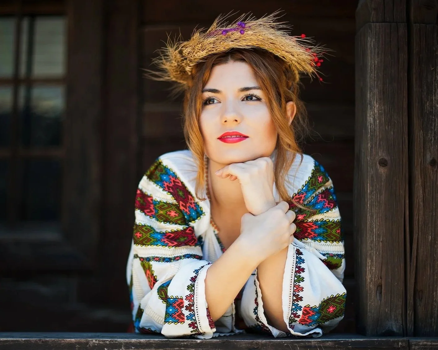 O femeie îmbrăcată în haine tradiționale brodate și cu o pălărie de paie împodobită cu flori, cu bărbia sprijinită în mână, privind gânditoare în afara unei clădiri rustice din lemn.