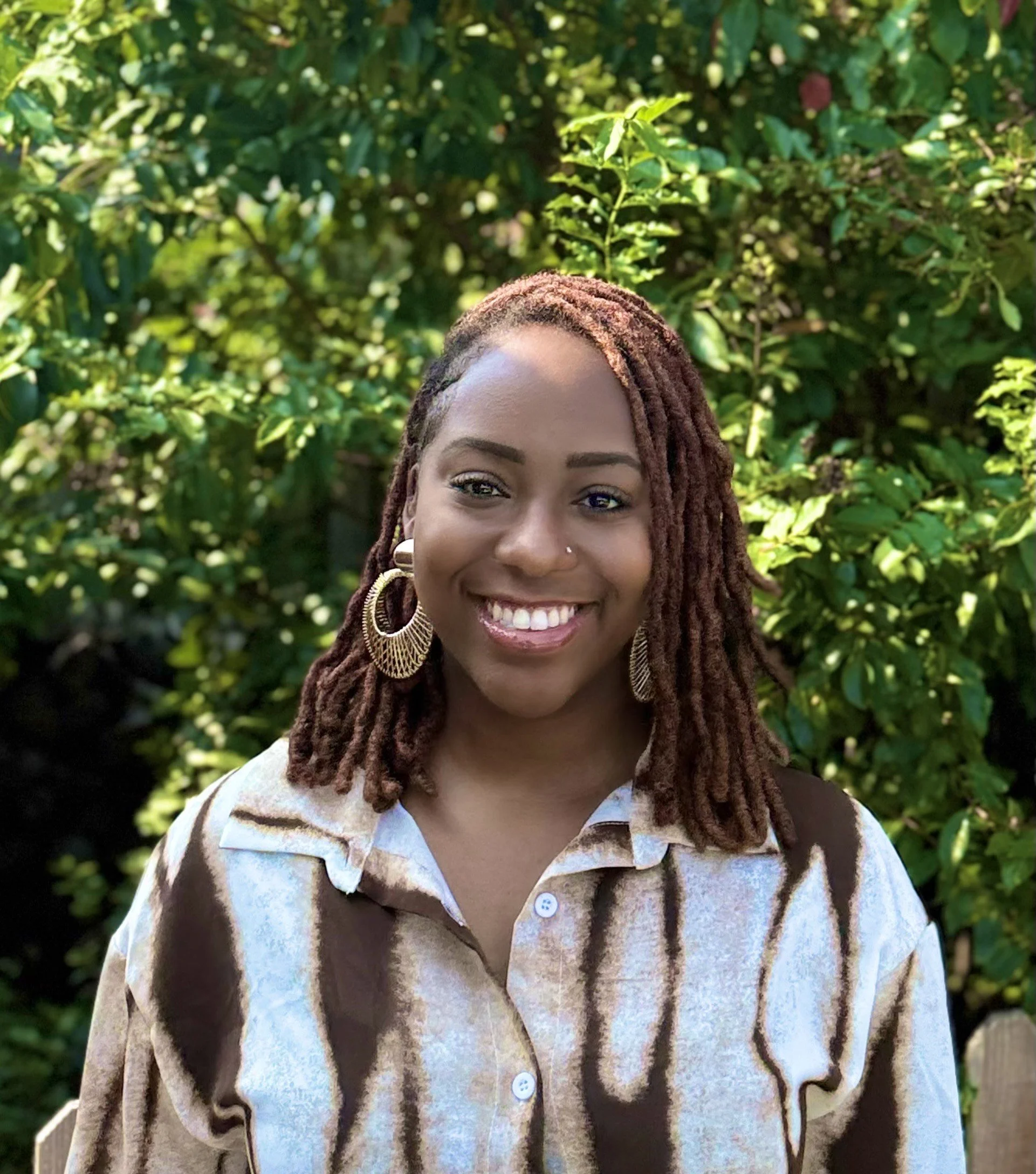 Close-up of a smiling woman with reddish-brown dreadlocks, gold hoop earrings, and a nose piercing, wearing a beige and brown striped button-up shirt, standing outdoors in front of green leafy bushes.