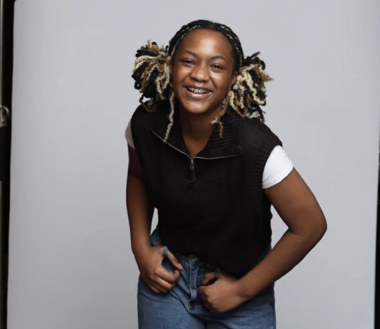 A young woman with dreadlocks smiling and posing with her hands on her hips in front of a plain white background.
