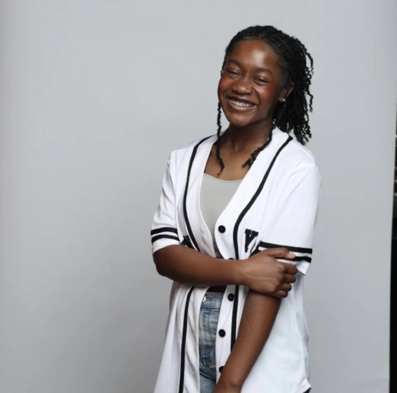 A smiling young woman with dark curly hair, wearing a baseball jersey-style shirt, standing against a plain light gray background.