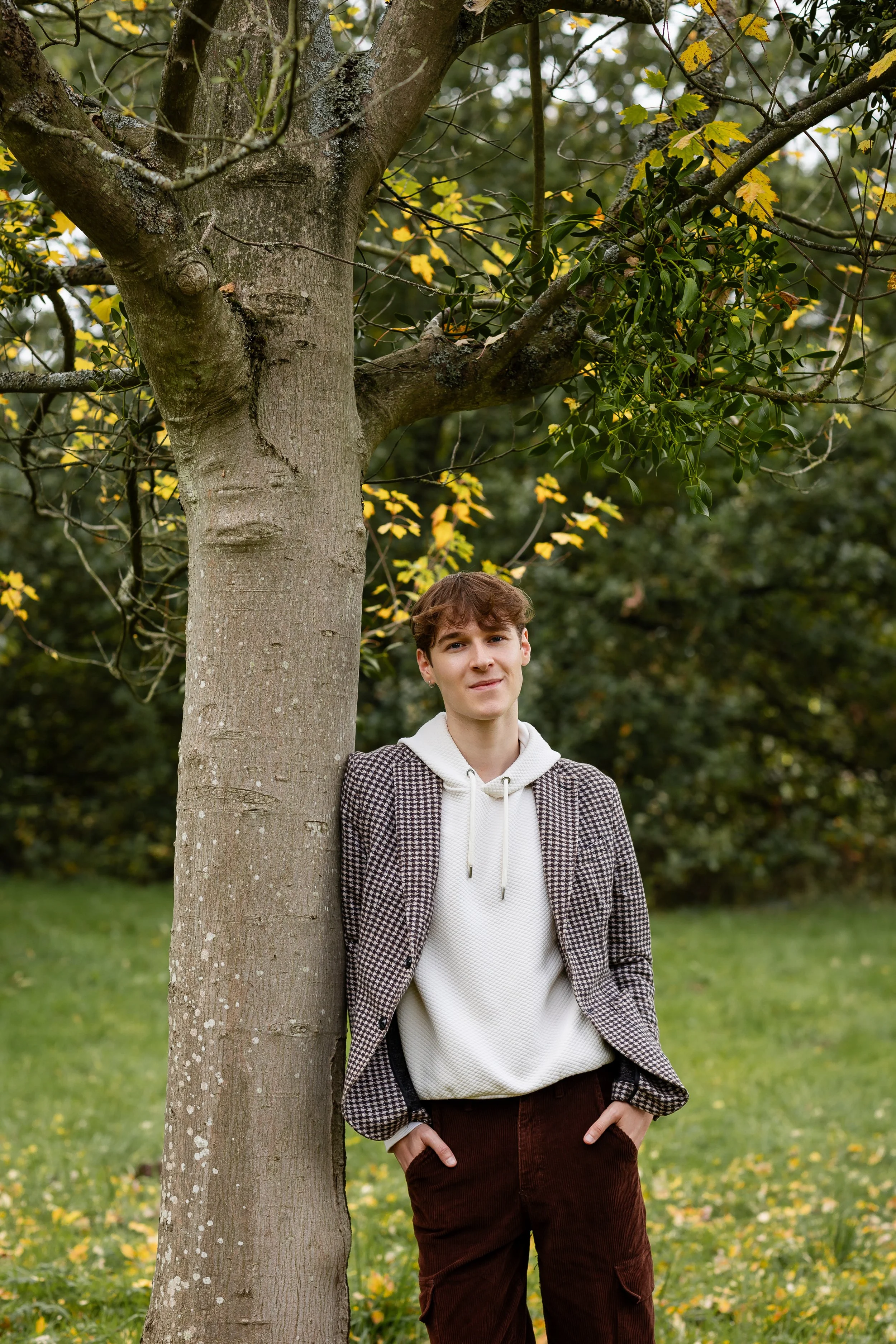 Me standing beside a tree with a mistletoe in a park, wearing a checked blazer, white hoodie, and brown pants.