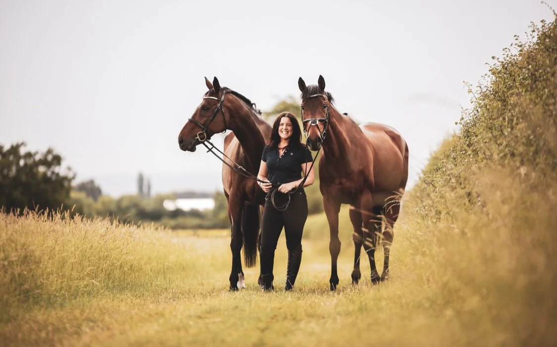 A woman standing between two brown horses on a grassy field, holding their reins, with trees and a cloudy sky in the background.